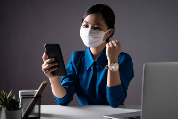 Donna asiatica in camicia blu con maschera protettiva che mostra un gesto vincente in ufficio. isolato su sfondo. Chiave bassa. - Foto, immagini