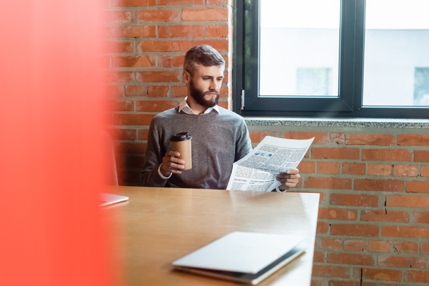 вибірковий фокус бородатого бізнесмена, який тримає каву, щоб піти і читати газету біля ноутбука
  - Фото, зображення