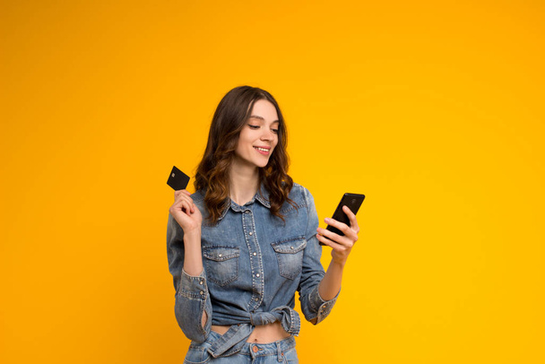Young pretty smiling brunette woman in denim shirt toothy smiling holding plastic credit card and looking at mobile phone screen,standing isolated on yellow background.Online shopping - Foto, immagini