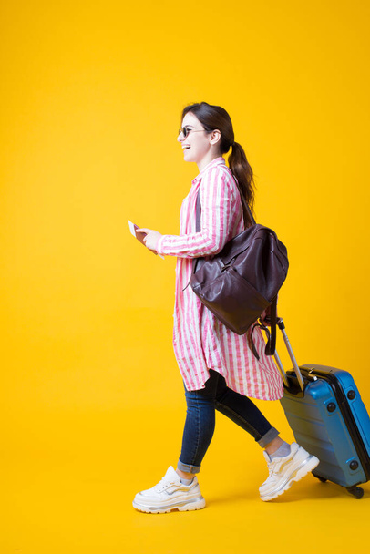 Захоплена стильна брюнетка мандрівник-туристична жінка в літньому одязі в білих кросівках з валізою ізольована на жовтому тлі. Щаслива жінка-пасажир перед польотом за кордон. вертикально
 - Фото, зображення