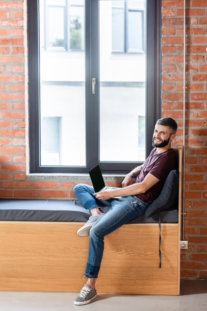hombre de negocios feliz usando el ordenador portátil con la pantalla en blanco mientras está sentado en el banco de la ventana en la oficina moderna
 - Foto, Imagen