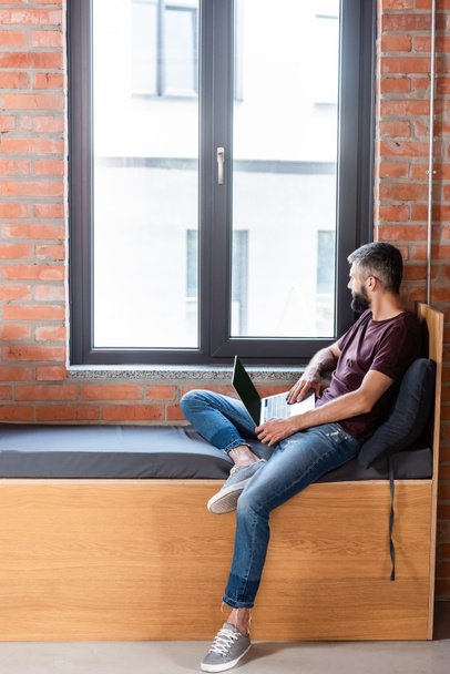 homem de negócios barbudo usando laptop com tela em branco enquanto sentado no banco de janela no escritório moderno
 - Foto, Imagem