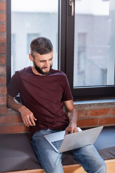 bearded businessman sitting on window bench and using modern laptop  - Фото, изображение