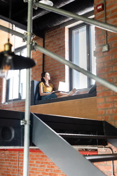 wybiórcze skupienie bizneswoman siedzi na ławce okien z laptopem i patrząc na okna w biurze  - Zdjęcie, obraz