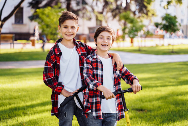 Twee aantrekkelijke Europese jongens broers, in rood en wit geruite flanellen shirts, staand op scooters in het park. Ze lachen, glimlachen, knuffelen en hebben plezier. Grote vingers omhoog steken. - Foto, afbeelding