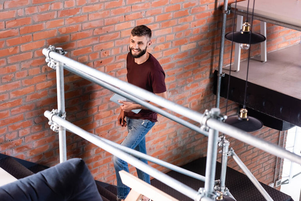 selective focus of cheerful businessman standing on stairs and holding laptop in modern office  - Photo, Image
