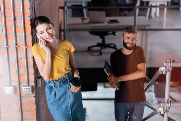 Селективный фокус счастливой деловой женщины, разговаривающей на смартфоне рядом с бородатым бизнесменом с ноутбуком
  - Фото, изображение