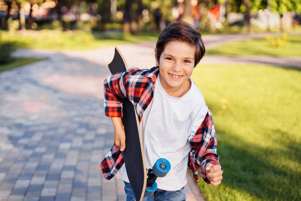 Happy young guy running along the alley on a sunny summer evening. Holding his skateboard in hands. Actively spending summer holidays. Dressed in a plaid shirt and white T-shirt. - Foto, imagen