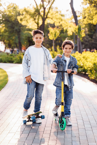 Aktivní dětská zábava v letním parku. Dva roztomilí bratři se baví v parku o letním večeru. Jízda na skateboardu a skútru. Oblečený v džínových oblecích a bílých tričkách. - Fotografie, Obrázek