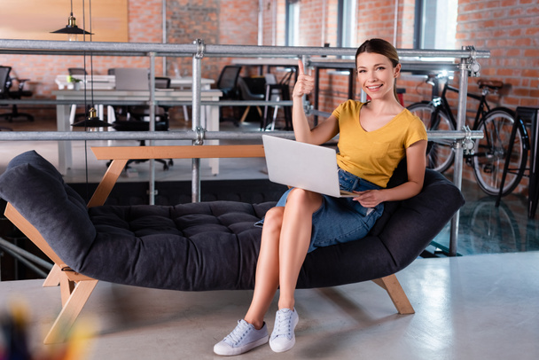 selektywne skupienie szczęśliwy businesswoman pokazując kciuk w górę podczas korzystania z laptopa i siedzi na kanapie w biurze  - Zdjęcie, obraz