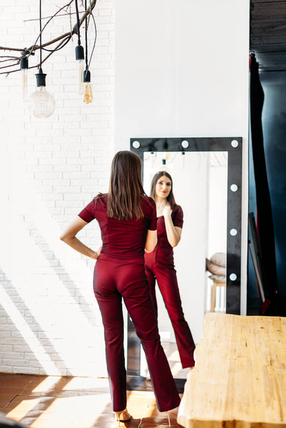 A girl in a red suit stands in front of a mirror, a large mirror, a reflection, trying on a suit, tailoring, outfit, going to a party or to a party - Фото, изображение
