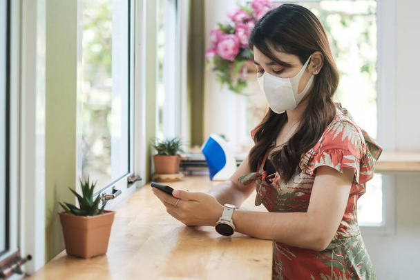 Aziatische vrouw dragen beschermende gezichtsmasker en met behulp van smartphone in restaurant, beschermen coronavirus flexie. sociale afstandelijkheid, nieuw normaal en leven na de covid-19 pandemie - Foto, afbeelding