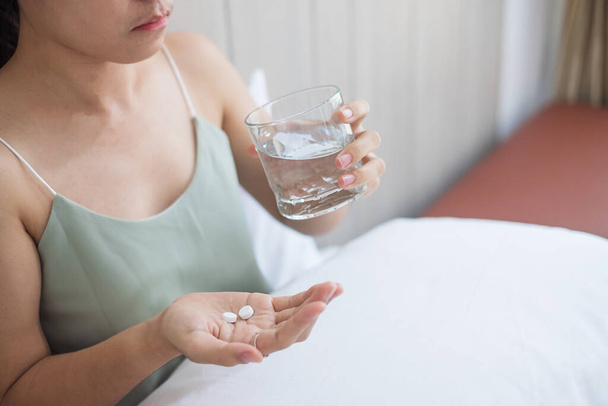 Взрослая женщина с таблетками и стаканом воды, принимающая лекарства на кровати утром дома. Мигрень, обезболивающее, головная боль, грипп, болезни, болезни и концепция здравоохранения
 - Фото, изображение