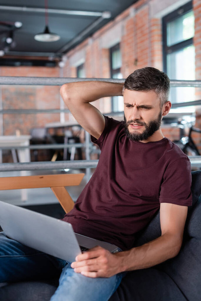 displeased businessman sitting on sofa and using laptop in office - Photo, Image