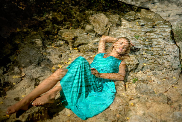 girl is drowned under water. blonde woman wearing a blue dress. sleep beauty charming sea princess resting on wet stones, girl in blue long tender dress lies in cold waterl hands under head hair, young female - Фото, изображение