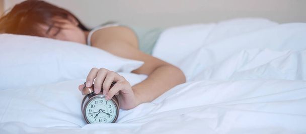 Wecker und asiatische Frauenhand stoppen die Zeit im Bett, während sie schlafen, junge erwachsene Frauen wachen spät am Morgen auf. Frisch entspannen, schläfrig sein und einen schönen Tag haben - Foto, Bild