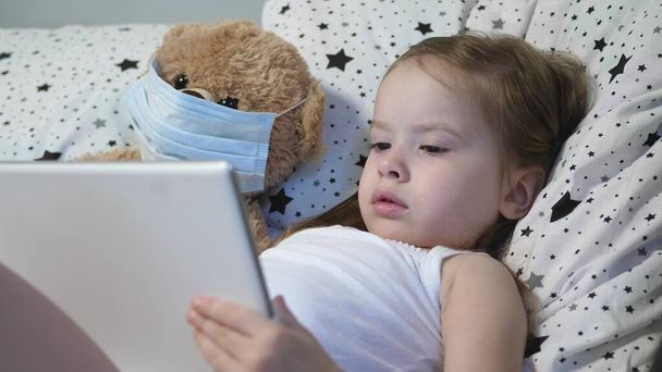 Маленький ребенок с медведем в маске играет на планшете, лежа на кровати. Ковид-19. Ребенок в карантине дома. Дистанционное обучение для дошкольников. Современные технологии - Фото, изображение