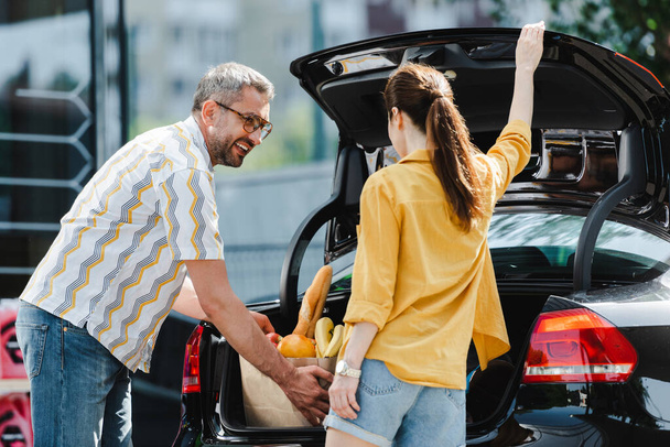 Selective focus of woman opening car trunk near smiling man holding shopping bag with food on urban street  - Photo, Image