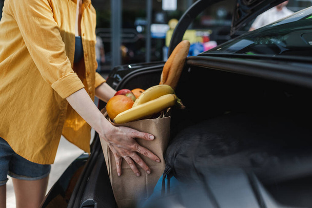 Обрезанный вид женщины положить сумку с едой в багажник автомобиля на городской улице
  - Фото, изображение