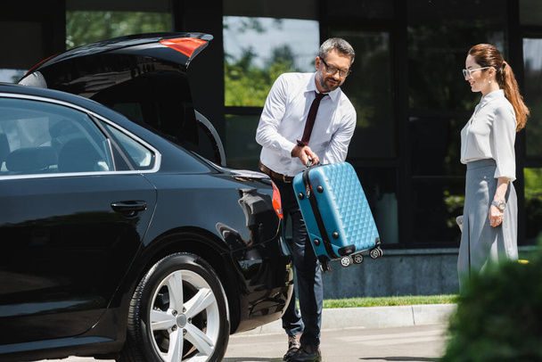 Selective focus of smiling businesswoman standing near colleague putting suitcase in car trunk on urban street  - Photo, Image