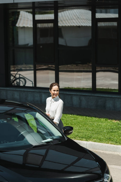 Mujer de negocios sonriente sosteniendo anteojos cerca de coche en la calle urbana
  - Foto, Imagen