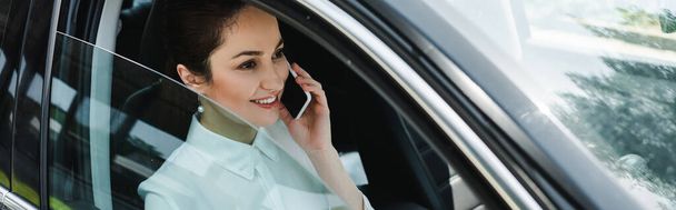 Panoramic crop of smiling businesswoman talking on smartphone in car  - Photo, Image