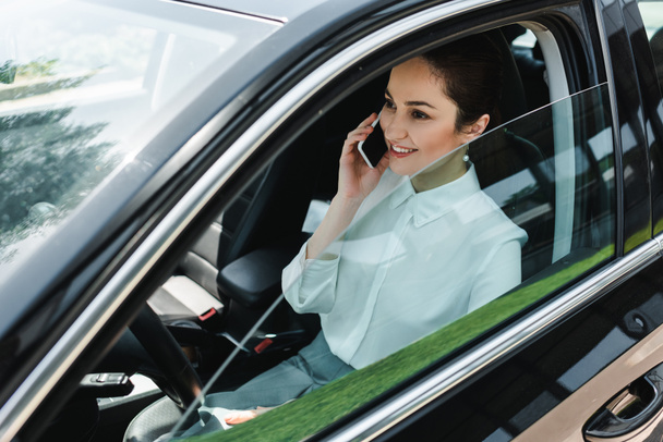 Lächelnde Geschäftsfrau spricht auf Smartphone, während sie auf dem Fahrersitz im Auto sitzt  - Foto, Bild