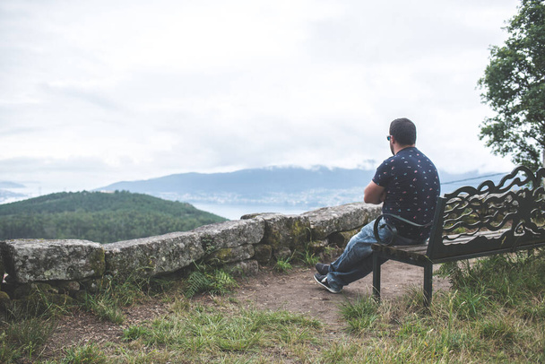 Homem sentado em um banco sozinho, contemplando uma paisagem nebulosa
 - Foto, Imagem
