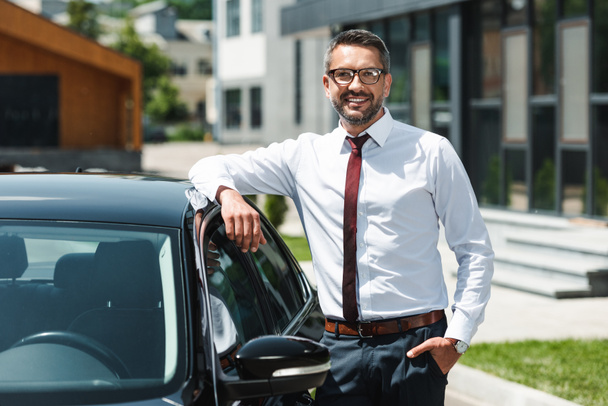 Selective focus of businessman with hand in pocket smiling at camera near car on urban street  - Fotografie, Obrázek