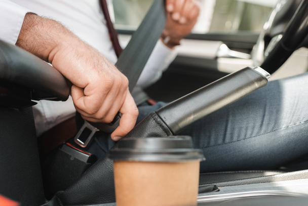 Selective focus of businessman fastening safety belt near paper cup in car  - Foto, imagen