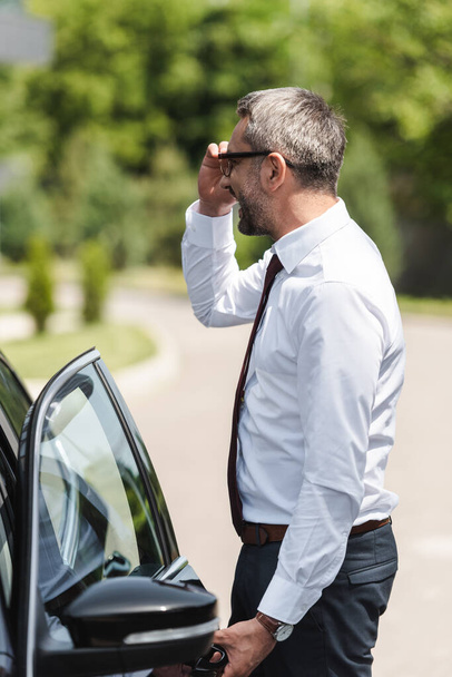 Vista lateral del empresario positivo ajustando las gafas y abriendo la puerta del coche en la calle urbana
  - Foto, imagen
