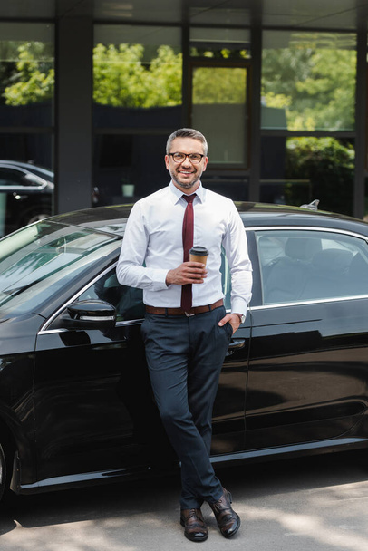 Successful businessman smiling at camera while holding cup of coffee near car on urban street  - Foto, imagen