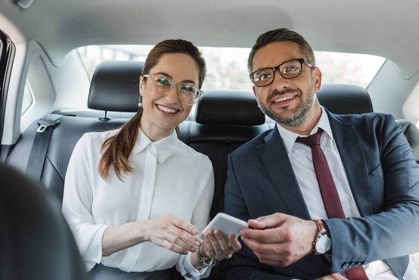 Selektivní zaměření usměvavé podnikatelky držící smartphone a dívající se na kameru v blízkosti podnikatele v autě  - Fotografie, Obrázek