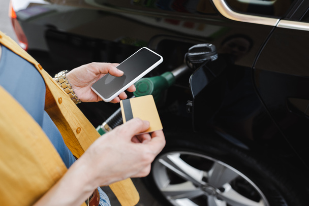 Vista cortada da mulher segurando smartphone e cartão de crédito enquanto reabastece o carro no posto de gasolina
  - Foto, Imagem