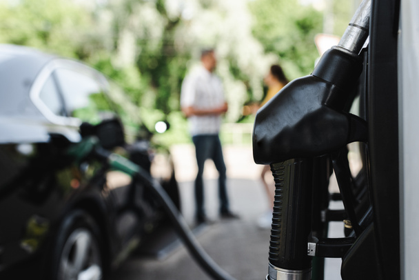 Επιλεκτική εστίαση του ακροφυσίου τροφοδοσίας σε βενζινάδικο και ζευγάρι στέκεται κοντά σε εξωτερικούς χώρους αυτοκινήτων  - Φωτογραφία, εικόνα