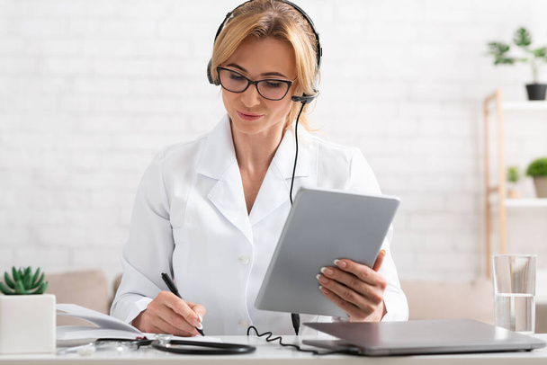 Female doctor makes notes in notebook, holds tablet in hand in interior of office - Фото, изображение