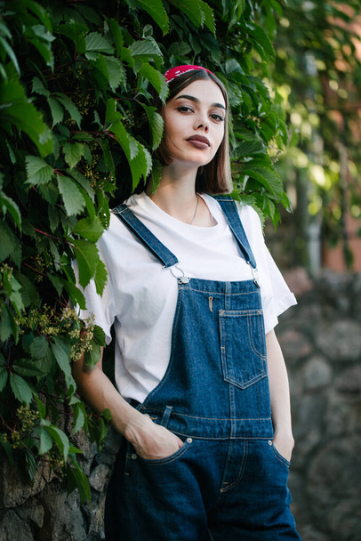 Slim sexy brunette girl wearing woman's denim overall over green tree wall texture in urban area - 写真・画像