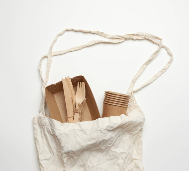 λευκή υφασμάτινη τσάντα και επιτραπέζια σκεύη μίας χρήσης από καφέ χαρτί χειροτεχνίας σε λευκό φόντο. Άποψη από ψηλά, έννοια της πλαστικής απόρριψης, μηδενικά απόβλητα  - Φωτογραφία, εικόνα