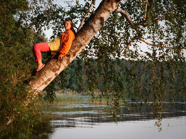 όμορφο κορίτσι σε ένα δέντρο πάνω από το νερό στο ηλιοβασίλεμα 2020 - Φωτογραφία, εικόνα
