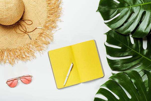 top view of green palm leaves, straw hat, sunglasses and yellow notepad on white background - Photo, Image