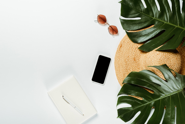 bovenaanzicht van groene palmbladeren, strohoed, zonnebril en notitieblok met smartphone op witte achtergrond - Foto, afbeelding