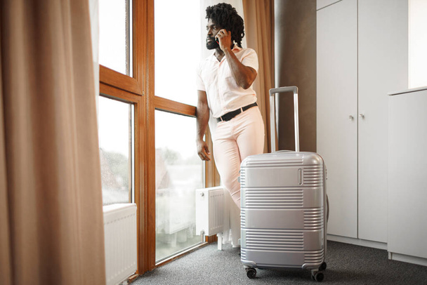 Portrait of a black man businessman with packed suitcase near large window in hotel room - Foto, imagen