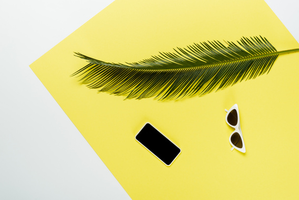 widok z góry zielony liść palmy w pobliżu okularów przeciwsłonecznych i smartfona na białym i żółtym tle - Zdjęcie, obraz