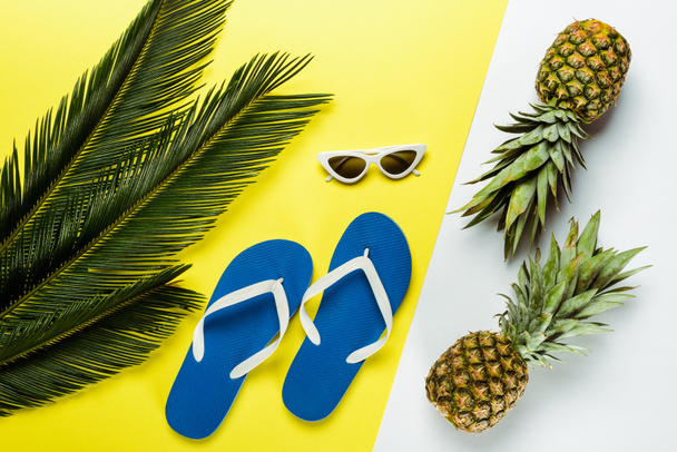 top view of green palm leaves, pineapples, sunglasses and blue flip flops on white and yellow background - Photo, Image