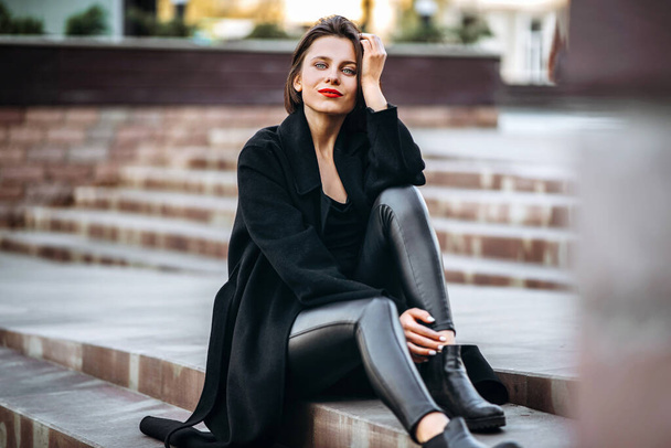 Full length portrait of young woman with short haircut and red lips, dressed in black. Woman sitting on the stairs outdoors on the stairs - Foto, imagen