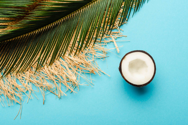 top view of green palm leaf, coconut half, straw hat on blue background - Zdjęcie, obraz