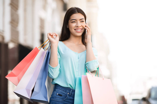 Вызываю такси. Радостная азиатская девушка держит в руках сумки с покупками, разговаривая по мобильному телефону на улице
 - Фото, изображение