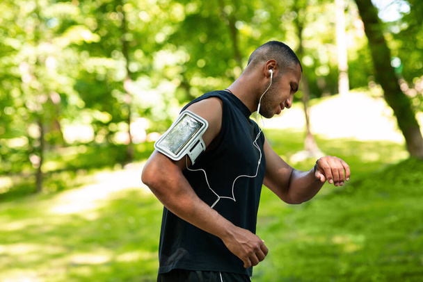 Вид сбоку афроамериканского бегуна, проверяющего смартфон или фитнес-трекер во время утренней пробежки в парке
 - Фото, изображение