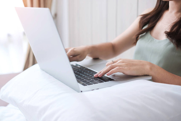 寝室の白い枕の上にコンピュータのラップトップを座って使用して幸せなカジュアルな女性。若い大人の女性朝の家庭やオンラインショッピングから働く。フリーランス、ライフスタイル、テクノロジーの概念 - 写真・画像