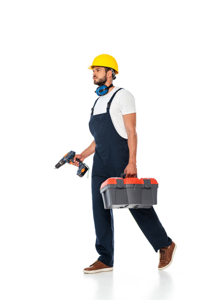 Handsome workman in overalls and hardhat holding toolbox and electric screwdriver while walking on white background - Photo, Image